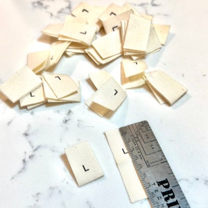 May include: A pile of white fabric labels with the letter "L" printed in black. The labels are approximately 1 inch by 1.5 inches in size. A ruler is visible in the bottom right corner of the image.
