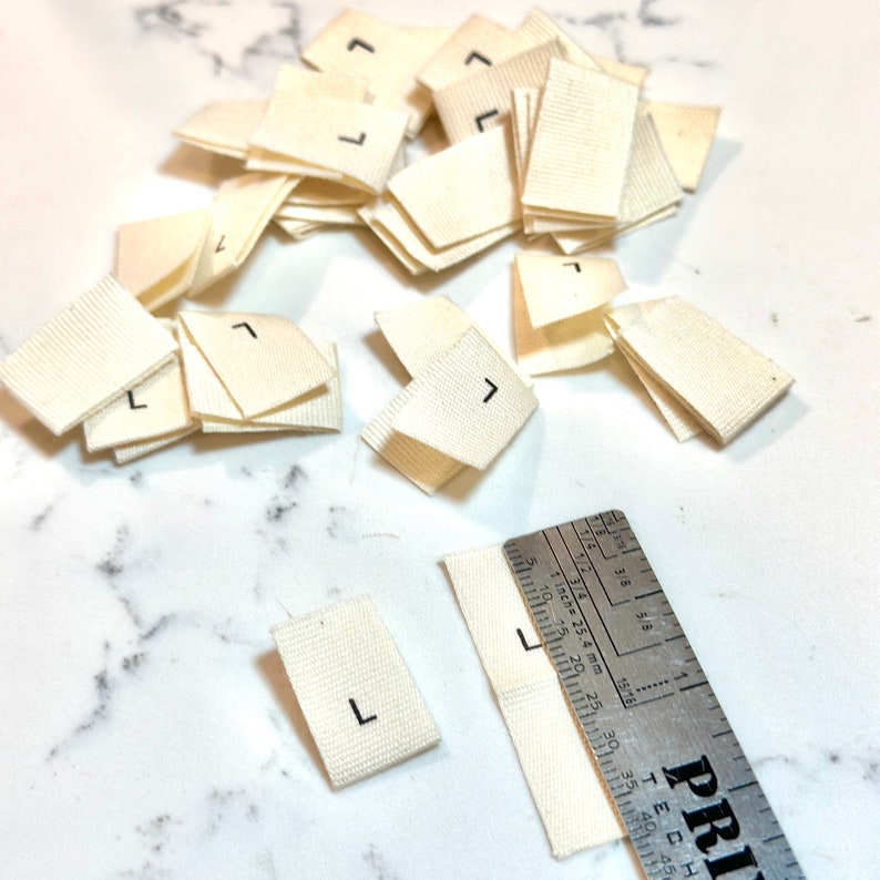 May include: A pile of white fabric labels with the letter "L" printed in black. The labels are approximately 1 inch by 1.5 inches in size. A ruler is visible in the bottom right corner of the image.