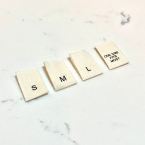 May include: Four white fabric labels with black text. The labels read "S", "M", "L", and "One Size Fits Most".