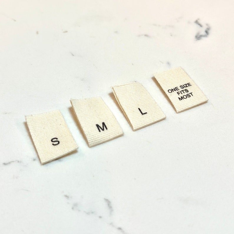May include: Four white fabric labels with black text. The labels read "S", "M", "L", and "One Size Fits Most".
