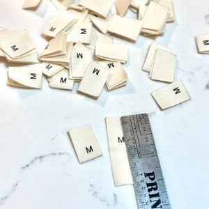 May include: A close-up of a pile of white fabric labels with the letter "M" printed on them. There are also a few labels with the letter "W" printed on them. A ruler is visible in the bottom right corner of the image.