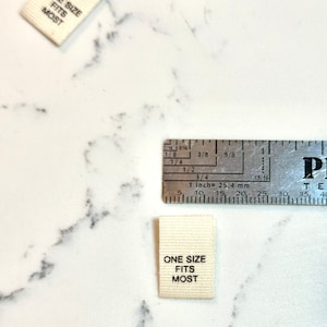 May include: A white fabric label with black text that reads "ONE SIZE FITS MOST". The label is measured with a ruler that shows 1 inch is equal to 25.4 mm.