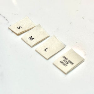May include: Four white fabric clothing size tags with black text. The tags read "S", "M", "L", and "One Size Fits Most".