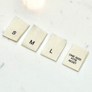 May include: Four white fabric tags with black text. The tags read "S", "M", "L", and "One Size Fits Most".