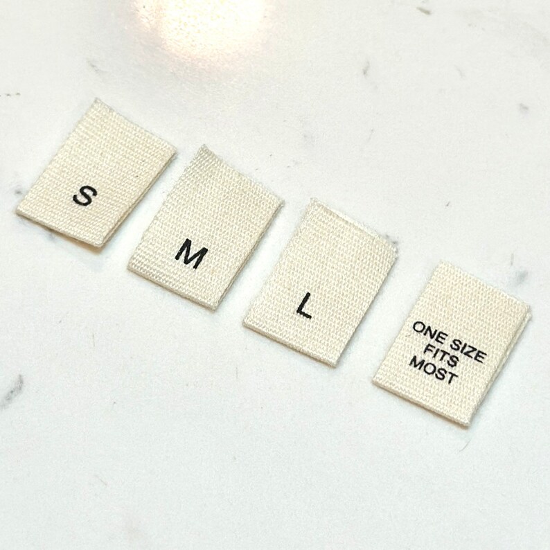 May include: Four white fabric tags with black text. The tags read "S", "M", "L", and "One Size Fits Most".
