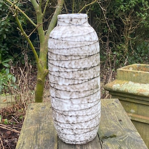 May include: A tall, white ceramic vase with a textured, striped pattern. The vase has a slightly rounded base and a narrow opening.
