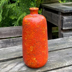 May include: A cylindrical ceramic vase with a narrow neck and a rounded rim. The exterior surface features a vibrant, textured orange glaze with mottled yellow and brown undertones, creating a rugged, uneven appearance across the entire body.