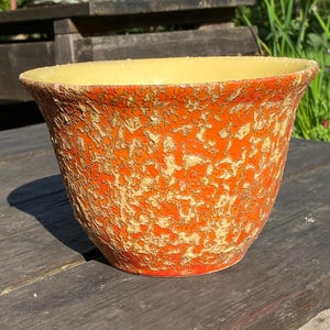 May include: A ceramic plant pot with a textured orange and cream exterior and a yellow interior. The pot has a wide, slightly flared rim. The pot is designed for indoor or outdoor use and sits on a wooden surface.