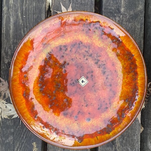 May include: A circular ceramic plate with a glossy, glazed finish. The plate displays a blend of orange, red, and brown tones, with a speckled texture. A small, square sticker is visible in the center. The plate's design is reminiscent of a sunset.