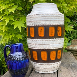 May include: A tall, cream-coloured ceramic vase with brown and orange rectangular details. A smaller, dark blue ceramic jug with a textured surface is next to the vase. Both are on a wooden surface.
