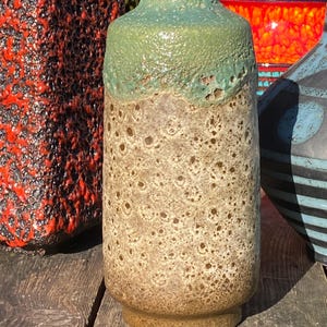 May include: A tall, cylindrical ceramic vase with a textured, speckled surface. The vase has a light brown base and body, topped with a textured, light green rim. Other colourful vases are visible in the background.