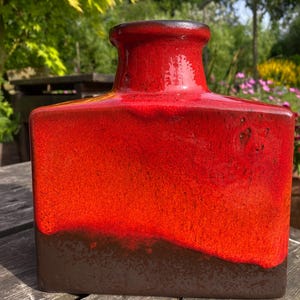 May include: A red ceramic vase with a dark brown base and rim. The vase has a rectangular shape and a glossy finish. The vase is a decorative item. The vase is sitting on a wooden surface. The vase is approximately 20 cm tall.