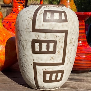 May include: A ceramic vase with a white, speckled, textured surface featuring a dark brown geometric pattern consisting of interconnected lines and rectangular shapes. The vase has a rounded body and a wide opening at the top, showcasing a distinct contrast between the light, mottled base and the dark, bold graphic design.