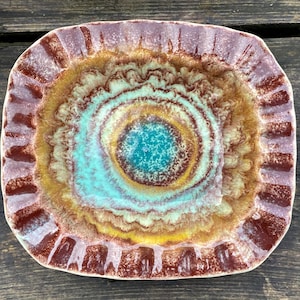 May include: A square ceramic plate with a distinctive, colourful design. The plate features concentric circles in shades of turquoise, yellow, and brown, with a textured rim in maroon and lavender. A decorative piece.