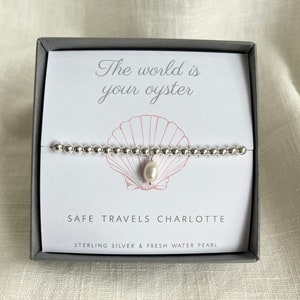 K&ouml;nnte beinhalten: Ein silbernes Armband mit einem Perlenanh&auml;nger in einer Schachtel, auf der "The world is your oyster" und "Safe Travels Charlotte" steht, sowie der Text "Sterling Silver & Fresh Water Pearl".