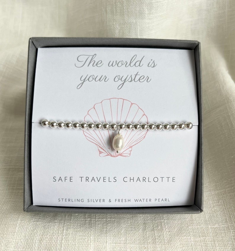 K&ouml;nnte beinhalten: Ein silbernes Armband mit einem Perlenanh&auml;nger in einer Schachtel, auf der "The world is your oyster" und "Safe Travels Charlotte" steht, sowie der Text "Sterling Silver & Fresh Water Pearl".