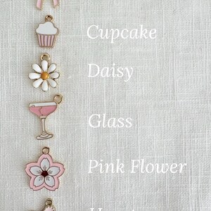 May include: A collection of pink and gold charms, including a bow, cupcake, daisy, glass, pink flower, and heart. Each charm is detailed with enamel and has a loop for attaching to jewelry or other crafts. Text labels each charm.
