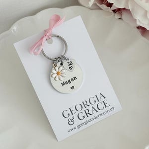 May include: A silver keychain with a daisy charm, a heart charm with the number 18, and a round charm with the name "Megan". The keychain is attached to a white card with the text "GEORGIA & GRACE" and a website address. A pink ribbon is tied to the top.