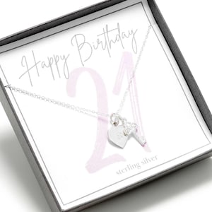 Op de afbeelding: Een zilveren ketting met een hart- en sleutelbedel in een cadeaudoos. De doos heeft een roze en wit ontwerp met de tekst "Happy Birthday 21" en "sterling silver".