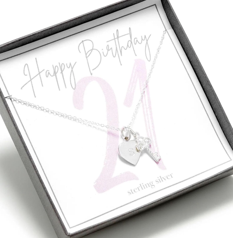 Op de afbeelding: Een zilveren ketting met een hart- en sleutelbedel in een cadeaudoos. De doos heeft een roze en wit ontwerp met de tekst "Happy Birthday 21" en "sterling silver".