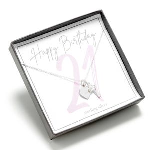 Op de afbeelding: Een zilveren ketting met een hartvormige hanger en een sleutel hanger. De ketting zit in een doos met een roze en witte achtergrond waarop "Happy Birthday 21" en "sterling silver" staat.