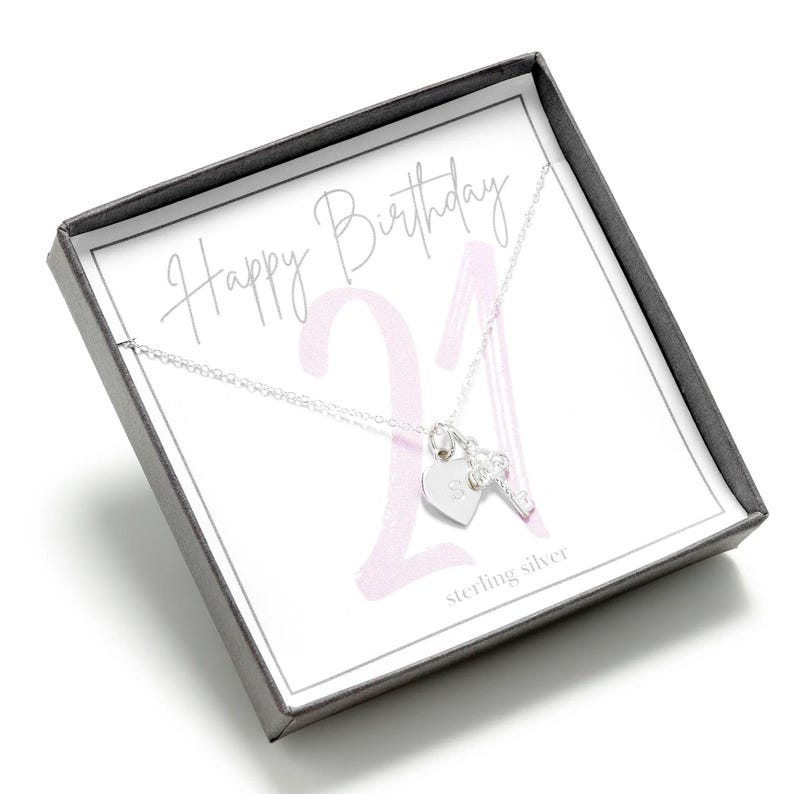 Op de afbeelding: Een zilveren ketting met een hartvormige hanger en een sleutel hanger. De ketting zit in een doos met een roze en witte achtergrond waarop "Happy Birthday 21" en "sterling silver" staat.
