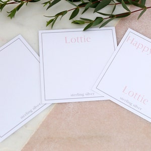 May include: Three white gift tags with black borders. The top tag says "Happy Birthday" in pink lettering. The middle tag says "Lottie" in pink lettering and "sterling silver" in black lettering. The bottom tag says "Lottie" in pink lettering and "sterling silver" in black lettering.