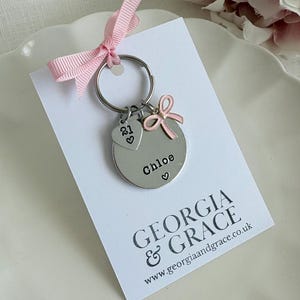 Pu&ograve; includere: Un portachiavi d'argento con un ciondolo rotondo che dice "Chloe" con un cuore. Il ciondolo ha anche un fiocco rosa e un piccolo ciondolo a forma di cuore con il numero "21" su di esso. Il portachiavi &egrave; su una carta bianca con il testo "GEORGIA & GRACE www.georgiaandgrace.co.uk"