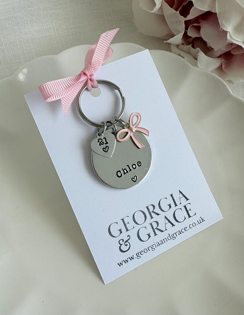 Pu&ograve; includere: Un portachiavi d'argento con un ciondolo rotondo che dice "Chloe" con un cuore. Il ciondolo ha anche un fiocco rosa e un piccolo ciondolo a forma di cuore con il numero "21" su di esso. Il portachiavi &egrave; su una carta bianca con il testo "GEORGIA & GRACE www.georgiaandgrace.co.uk"
