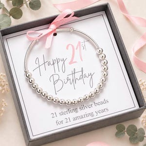 May include: A circular bracelet made of polished silver beads, presented in a dark gray gift box. The card inside features the text Happy 21st Birthday and 21 sterling silver beads for 21 amazing years, accented with a light pink ribbon bow.