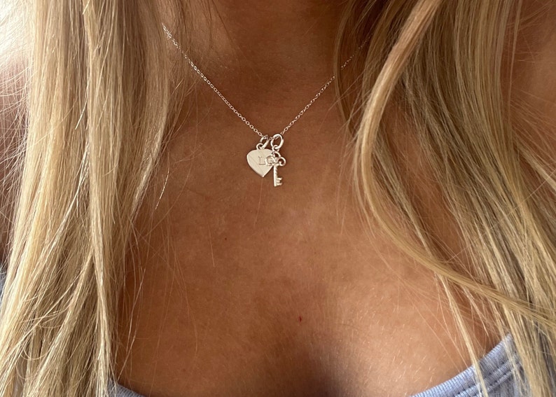 Op de afbeelding: Een zilveren ketting met een hartvormige hanger en een sleutel hanger. De hartvormige hanger heeft de letter "L" gegraveerd.