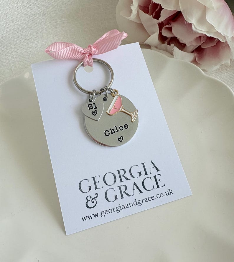 Pu&ograve; includere: Un portachiavi d'argento con un ciondolo a forma di cocktail rosa, un ciondolo a forma di cuore con il numero 21 e un'etichetta rotonda che dice "Chloe" con un cuore. Il portachiavi &egrave; su una carta bianca con il testo "GEORGIA & GRACE www.georgiaandgrace.co.uk"