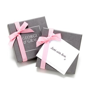 Op de afbeelding: Twee grijze cadeaudozen met roze strikken. De bovenste doos heeft een wit label met de tekst "GEORGIA & GRACE". De onderste doos heeft een witte kaart met de tekst "Sent with love" en een handgeschreven handtekening.