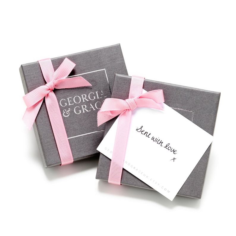 Op de afbeelding: Twee grijze cadeaudozen met roze strikken. De bovenste doos heeft een wit label met de tekst "GEORGIA & GRACE". De onderste doos heeft een witte kaart met de tekst "Sent with love" en een handgeschreven handtekening.