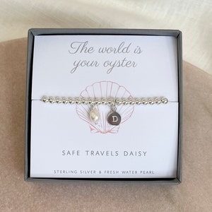 K&ouml;nnte beinhalten: Ein silbernes Armband mit einem Perlenanh&auml;nger und einem silbernen Anh&auml;nger mit dem Buchstaben "D" darauf. Das Armband befindet sich in einer Schachtel mit dem Text "Die Welt ist deine Auster" und "Safe Travels Daisy" darauf.