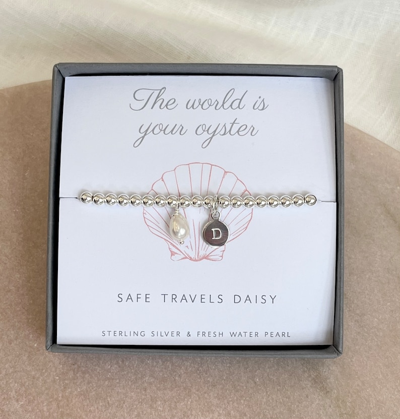 K&ouml;nnte beinhalten: Ein silbernes Armband mit einem Perlenanh&auml;nger und einem silbernen Anh&auml;nger mit dem Buchstaben "D" darauf. Das Armband befindet sich in einer Schachtel mit dem Text "Die Welt ist deine Auster" und "Safe Travels Daisy" darauf.