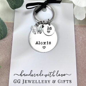以下が含まれることがあります： 蝶のチャーム、18とハートの丸いチャーム、そして「Alexis」とハートの大きな丸いチャームが付いたシルバーのキーホルダー。キーホルダーは、「handmade with love GG JEWELLERY & GIFTS」と書かれた白いカードの上に置かれています。