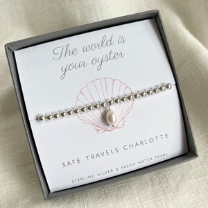 K&ouml;nnte beinhalten: Ein silbernes Armband mit einem S&uuml;&szlig;wasserperlen-Anh&auml;nger in einer grauen Box. Die Box tr&auml;gt den Text "The world is your oyster" und "Safe travels Charlotte" mit den Worten "Sterling Silver & Fresh Water Pearl" darunter.