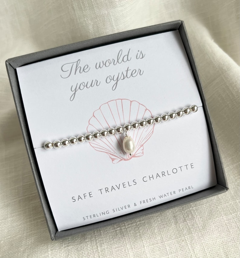 K&ouml;nnte beinhalten: Ein silbernes Armband mit einem S&uuml;&szlig;wasserperlen-Anh&auml;nger in einer grauen Box. Die Box tr&auml;gt den Text "The world is your oyster" und "Safe travels Charlotte" mit den Worten "Sterling Silver & Fresh Water Pearl" darunter.