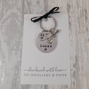 May include: Silver keychain with a round charm that says "Lucas" and "21" with a star. A small silver guitar charm is attached to the keychain. The keychain is on a white card with the text "handmade with love GG JEWELLERY & GIFTS".