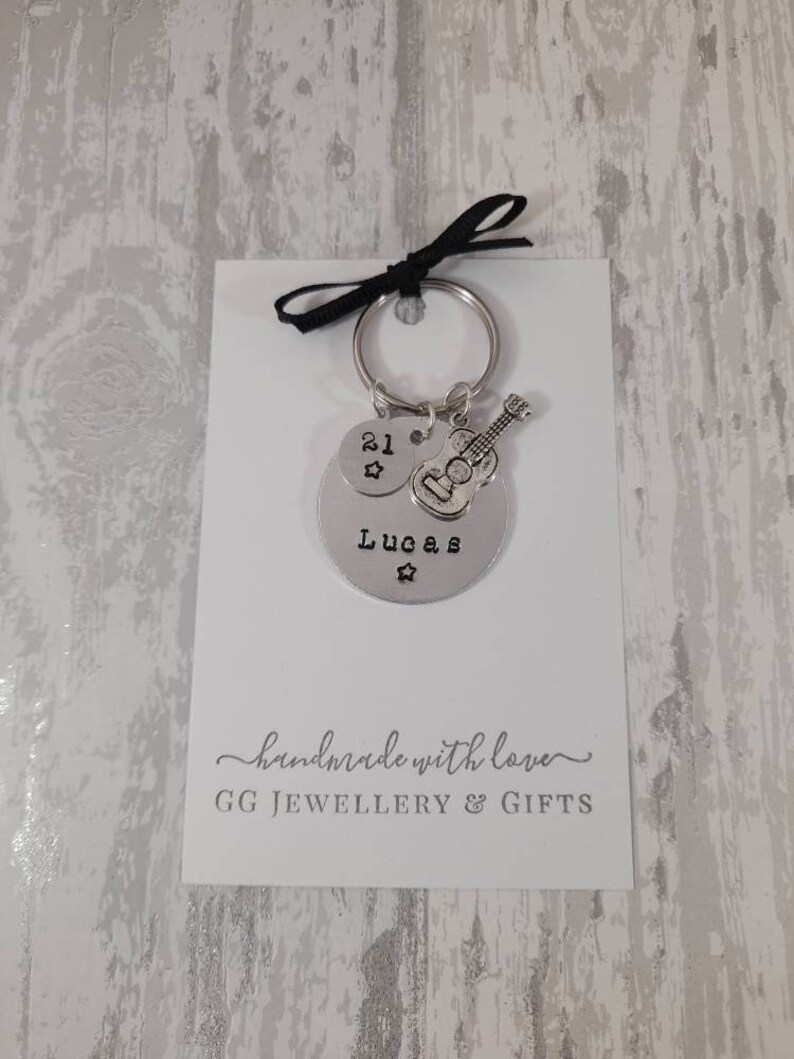May include: Silver keychain with a round charm that says "Lucas" and "21" with a star. A small silver guitar charm is attached to the keychain. The keychain is on a white card with the text "handmade with love GG JEWELLERY & GIFTS".