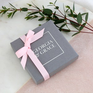 K&ouml;nnte beinhalten: Eine graue Geschenkbox mit einem rosa Band und dem Text "GEORGIA & GRACE" auf der Oberseite.