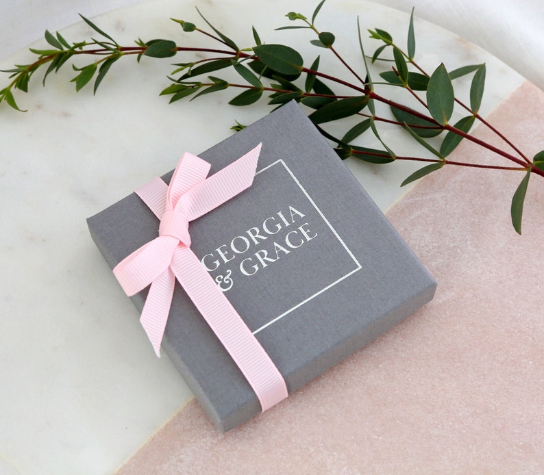 K&ouml;nnte beinhalten: Eine graue Geschenkbox mit einem rosa Band und dem Text "GEORGIA & GRACE" auf der Oberseite.