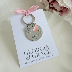 Pu&ograve; includere: Un portachiavi argentato con un ciondolo a forma di cocktail rosa, un ciondolo a forma di cuore con il numero 21 e un'etichetta rotonda che dice "Chloe" con un cuore. Il portachiavi &egrave; su una carta bianca con il testo "GEORGIA & GRACE" e "www.georgiaandgrace.co.uk".