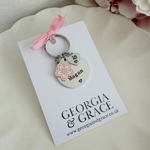 May include: A silver keychain with a pink ribbon, a round charm with the name "Megan" and a pink flower charm. The keychain also has a heart charm with the number "18". The card below reads "GEORGIA & GRACE".