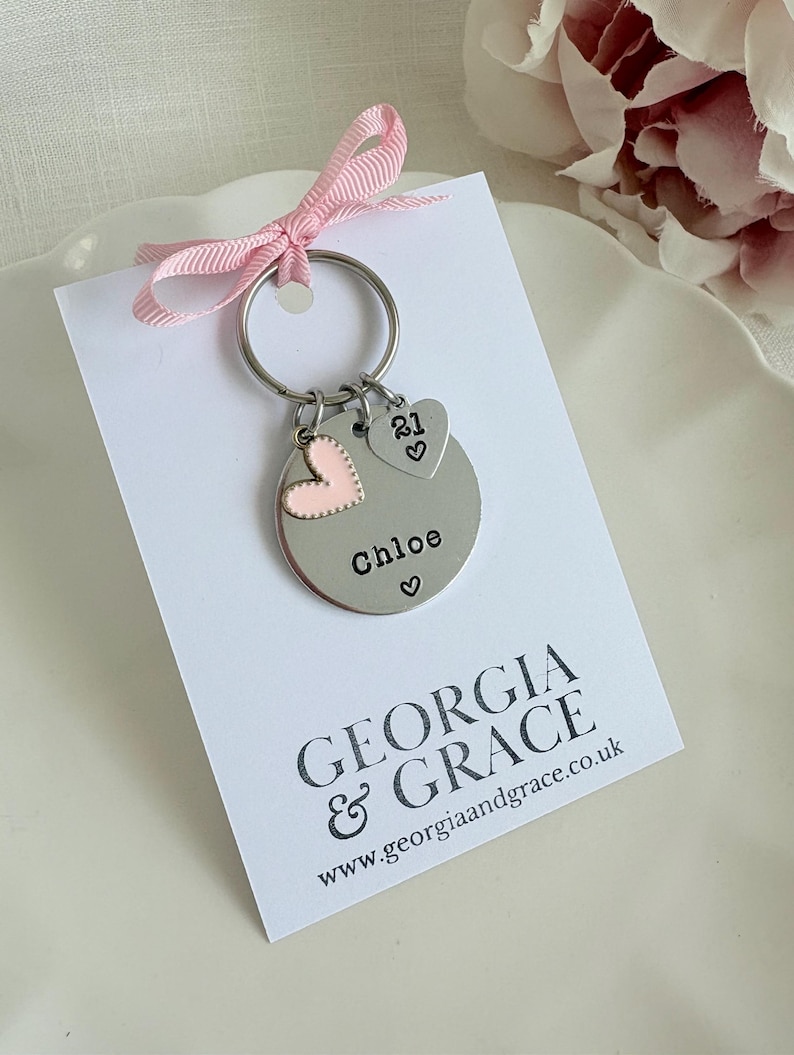 Pu&ograve; includere: Un portachiavi d'argento con un ciondolo a forma di cuore e un ciondolo rotondo che dice "Chloe" con un cuore. Il portachiavi &egrave; su una carta bianca con il testo "GEORGIA & GRACE www.georgiaandgrace.co.uk"