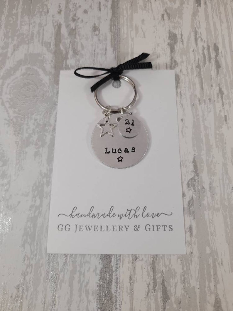 May include: Silver keychain with a star charm and a round tag that says "Lucas" and "21" with a star. The keychain is on a white card with the text "handmade with love" and "GG JEWELLERY & GIFTS".