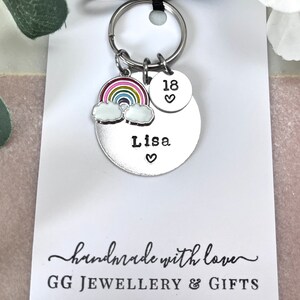 以下が含まれることがあります： 虹のチャーム、雲のチャーム、そして「Lisa」という名前と「18」という数字が刻まれた銀色の円盤が付いた銀色のキーホルダー。キーホルダーは、「手作りで愛情込めて」と「GG JEWELLERY & GIFTS」と書かれた白いカードの上に置かれています。