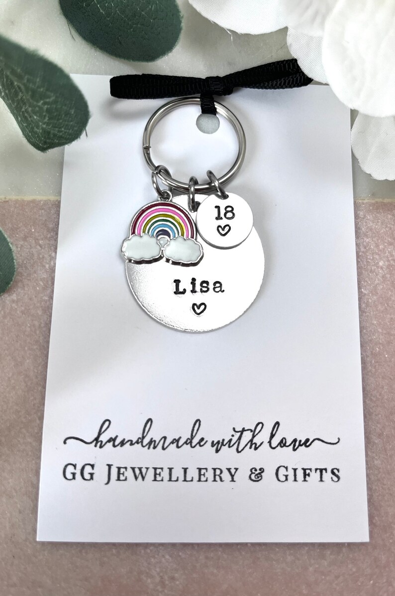 以下が含まれることがあります： 虹のチャーム、雲のチャーム、そして「Lisa」という名前と「18」という数字が刻まれた銀色の円盤が付いた銀色のキーホルダー。キーホルダーは、「手作りで愛情込めて」と「GG JEWELLERY & GIFTS」と書かれた白いカードの上に置かれています。
