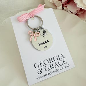 May include: A silver keychain with a pink bow and a charm that says "Megan" with a heart symbol. The keychain also has a heart-shaped charm with the number "18" and a pink ribbon. The card below the keychain has the text "GEORGIA & GRACE".